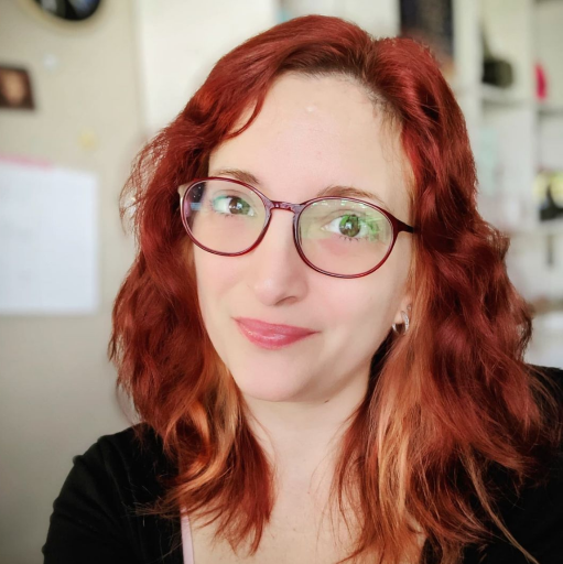 A small headshot of Leslie Salas. A hispanic white-passing woman with bright red hair and red-framed glasses. She has an amused, closed-lipped smile.
